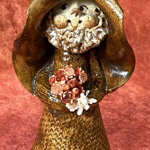 May include: A ceramic figurine of a gnome wearing a brown hat and holding a bouquet of dried flowers. The gnome is wearing a brown robe with a textured surface.