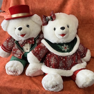 Könnte beinhalten: Zwei weiße Plüschbären, die in festliche Weihnachtskleidung gekleidet sind. Ein Bär trägt einen roten Samtanzug mit goldenen Verzierungen und einen Zylinder. Der andere Bär trägt ein rotes Samtkleid mit einem grünen Samtkragen und einem Schneeflockenmuster. Beide Bären haben das Datum 2003 auf ihren Pfoten.