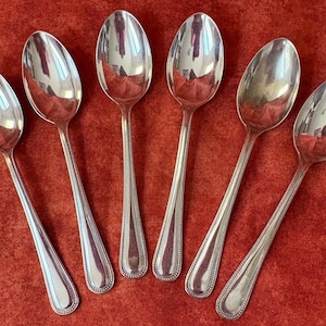 May include: Six silver spoons arranged on a red surface. The spoons have a classic design with a rounded bowl and a detailed handle. The spoons are reflective, showing the surrounding environment.
