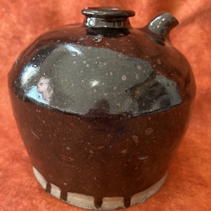May include: A dark brown ceramic jug with a speckled glaze and a white rim. The jug has a rounded body and a spout on the side.