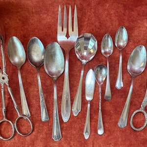 May include: An assortment of antique silver serving utensils, including spoons, a fork, and tongs, arranged on a textured red surface. The silverware features various designs and sizes, reflecting a vintage aesthetic.
