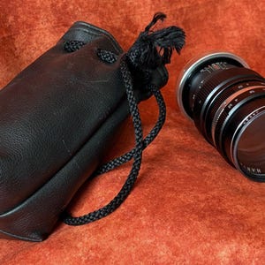 May include: A black leather camera lens case with a drawstring closure and a black camera lens. The lens has silver and black details and is lying on an orange surface.