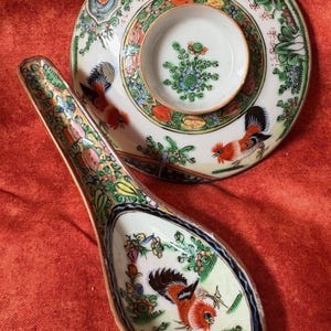 May include: A decorative ceramic spoon and small dish set. The spoon and dish are white with colorful floral and rooster designs. The spoon is long and curved, and the dish has a small, central well. The set is on a red surface.