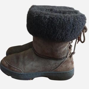May include: Brown suede boots with a black fur-like trim at the top. The boots have a round toe and a rubber sole. The back of the boots have a lace-up design. The boots are designed for cold weather.