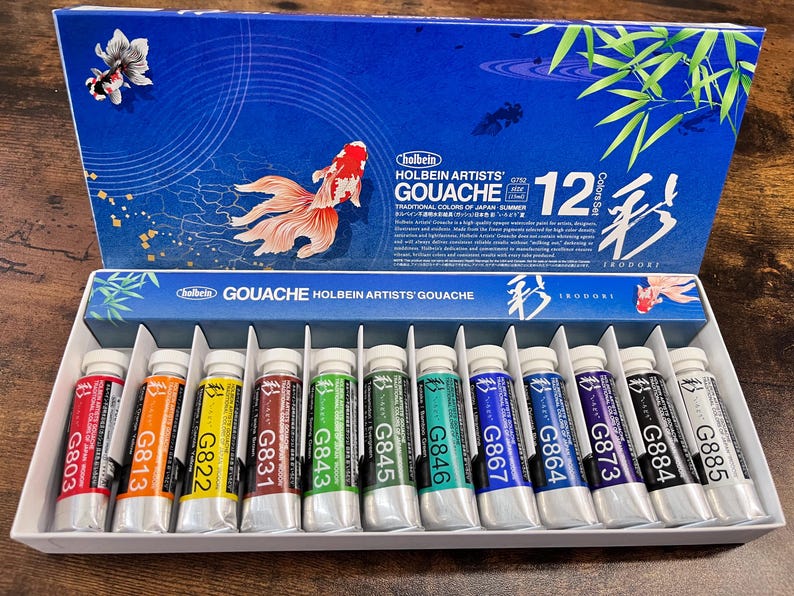 May include: A set of 12 Holbein Artists' Gouache paints in tubes. The tubes are labeled with the color number and name. The set is packaged in a blue box with a Japanese-inspired design. The box features a gold fish and the text "Holbein Artists' Gouache 12 Color Set Irodori."