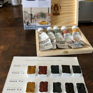 May include: A set of five watercolor paint tubes in a wooden box. The tubes are labeled "Horadam Aquarell" and are arranged in a row. The box is made of light wood and has a hinged lid. The paint tubes are a variety of colors, including yellow, red, green, brown, and gray.