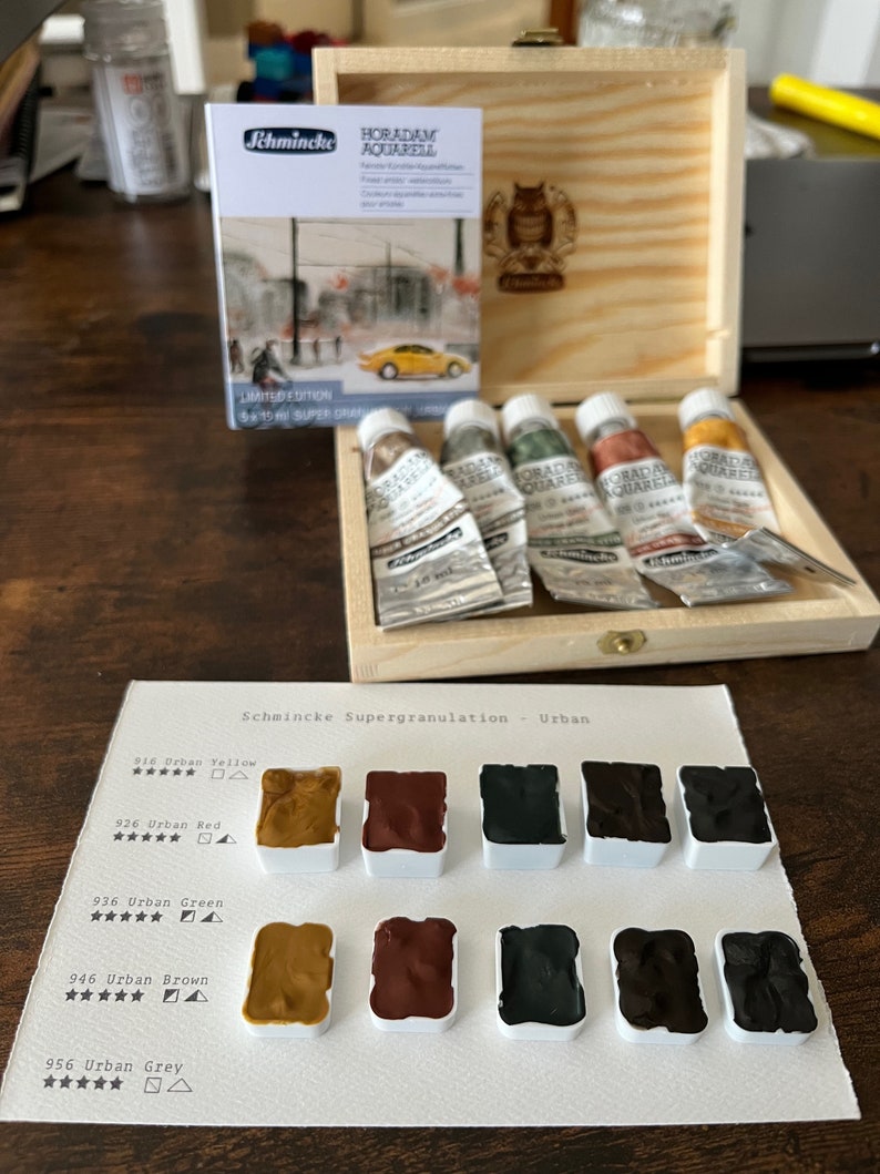 May include: A set of five watercolor paint tubes in a wooden box. The tubes are labeled "Horadam Aquarell" and are arranged in a row. The box is made of light wood and has a hinged lid. The paint tubes are a variety of colors, including yellow, red, green, brown, and gray.