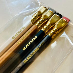 May include: Four pencils with gold-colored metal tips and erasers. The pencils are labeled "Blackwing" and "Blackwing 602".