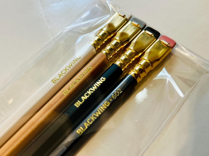May include: Four pencils with gold-colored metal tips and erasers. The pencils are labeled "Blackwing" and "Blackwing 602".