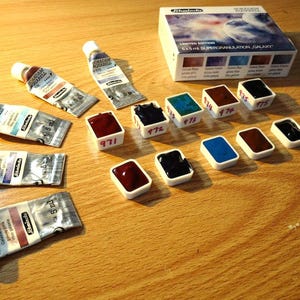 May include: A set of 12 watercolor paints in a variety of colors, including blue, green, brown, and black. The paints are in small, white pans with numbers printed on them. The paints are arranged in two rows on a wooden surface. The paint set is labeled "Limited Edition 5 ml Super Granulation Galaxy".