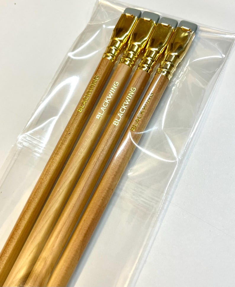 May include: Four wooden pencils with gold-colored tops and gray erasers. The pencils are packaged in a clear plastic bag. The pencils are labeled "Blackwing" in gold lettering.