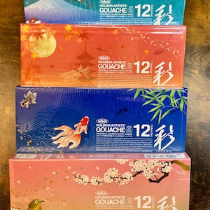 May include: Four boxes of Holbein Artists' Gouache paint, each featuring a different Japanese-inspired design. The boxes are stacked on top of each other, with the top box showing a pink and white cherry blossom design. The boxes are labeled with the words "Holbein Artists' Gouache" and "12 Color Set".