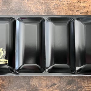 May include: A black rectangular paint palette with four compartments. The palette has a glossy finish and a gold emblem with the word "Schmincke" printed on the left side.