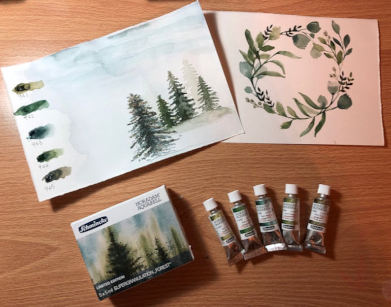 May include: A set of five 5 ml tubes of green watercolor paint, labeled "HORADAM Aquarell" and "Schmincke", with a box of watercolor paints labeled "Limited Edition 5x5ml Supergranulation - Forest". The box shows a watercolor painting of a forest scene. There are two watercolor paintings on paper, one of a forest scene and one of a green wreath.