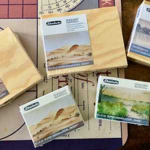 May include: Five wooden boxes of Horadam Aquarell watercolors. Each box features a different watercolor painting on the front. The boxes are labeled with the name of the watercolor and the size of the paint pans. The boxes are arranged in a row on a wooden surface.