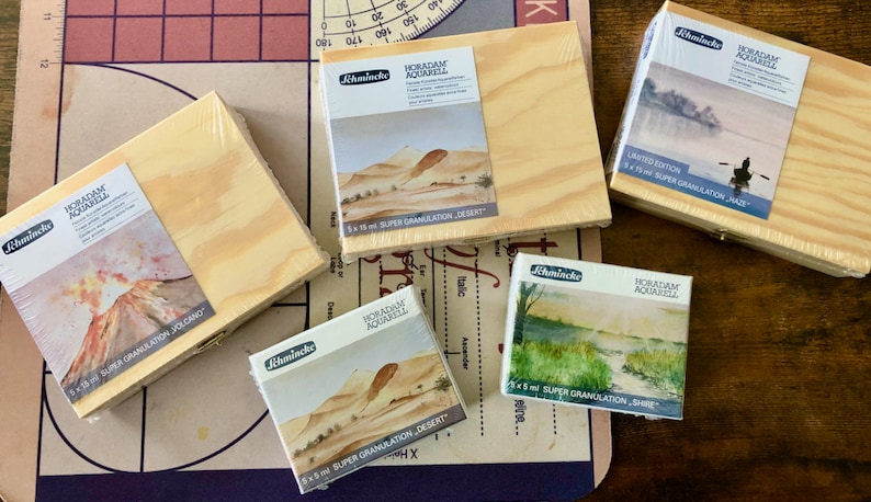 May include: Five wooden boxes of Horadam Aquarell watercolors. Each box features a different watercolor painting on the front. The boxes are labeled with the name of the watercolor and the size of the paint pans. The boxes are arranged in a row on a wooden surface.