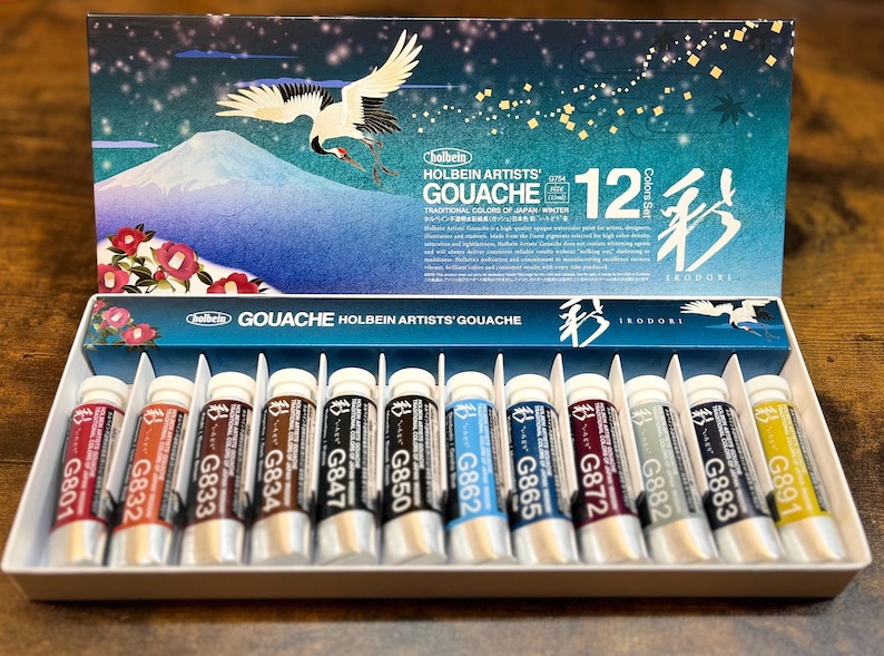 May include: A set of 12 Holbein Artists' Gouache paints in tubes. The tubes are labeled with the color number and name. The set is packaged in a box with a Japanese-inspired design featuring a crane and a mountain.