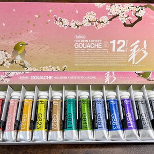 May include: A set of 12 gouache paints in tubes, arranged in a row. The tubes are labeled with the color number and name. The set is packaged in a box with a floral design and the text "Holbein Artists' Gouache 12 Colors Set".