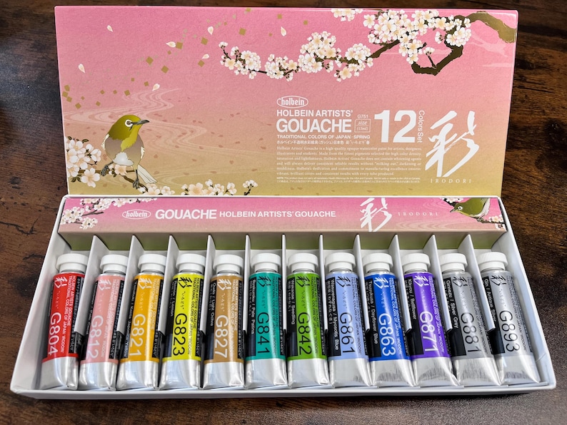 May include: A set of 12 gouache paints in tubes, arranged in a row. The tubes are labeled with the color number and name. The set is packaged in a box with a floral design and the text "Holbein Artists' Gouache 12 Colors Set".