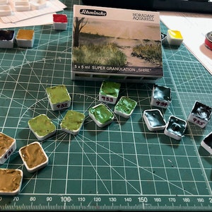 May include: A set of 10 watercolor paint pans in various shades of green and black. The pans are labeled with numbers, including 932, 933, 934, and 935. The pans are arranged on a green cutting mat with a ruler in the bottom left corner. The paint pans are labeled "Horadam Aquarell" and "Super Granulation Shire".