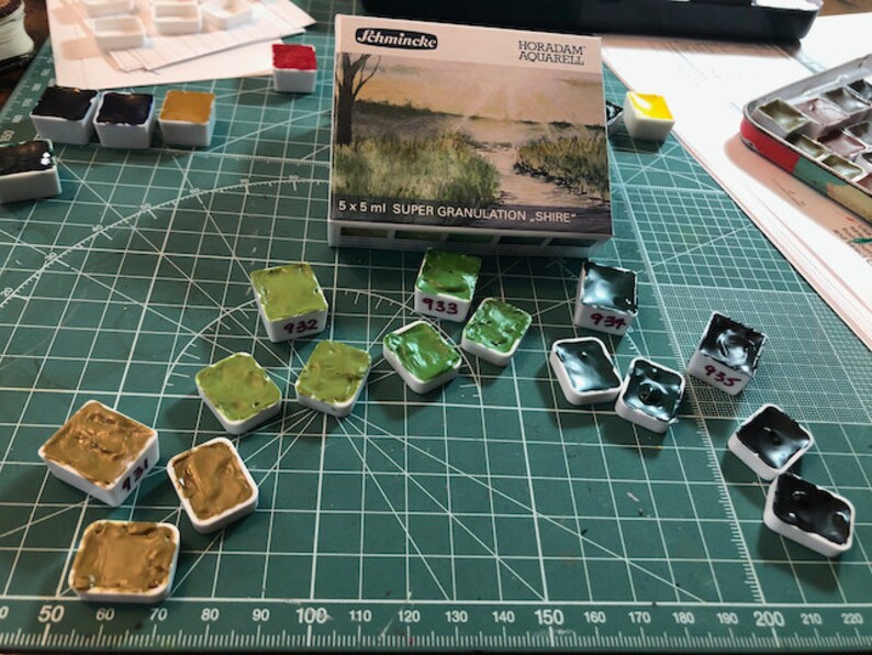 May include: A set of 10 watercolor paint pans in various shades of green and black. The pans are labeled with numbers, including 932, 933, 934, and 935. The pans are arranged on a green cutting mat with a ruler in the bottom left corner. The paint pans are labeled "Horadam Aquarell" and "Super Granulation Shire".