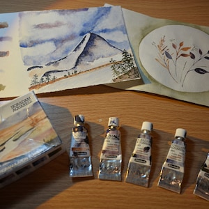 May include: A set of watercolor paints in tubes, arranged on a wooden surface. The tubes are labeled "HORADAM Aquarell" and are in shades of brown, blue, and green. There are also two watercolor paintings on paper, one depicting a mountain landscape and the other a floral design.