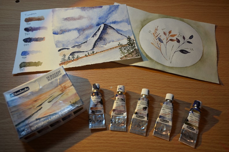 May include: A set of watercolor paints in tubes, arranged on a wooden surface. The tubes are labeled "HORADAM Aquarell" and are in shades of brown, blue, and green. There are also two watercolor paintings on paper, one depicting a mountain landscape and the other a floral design.