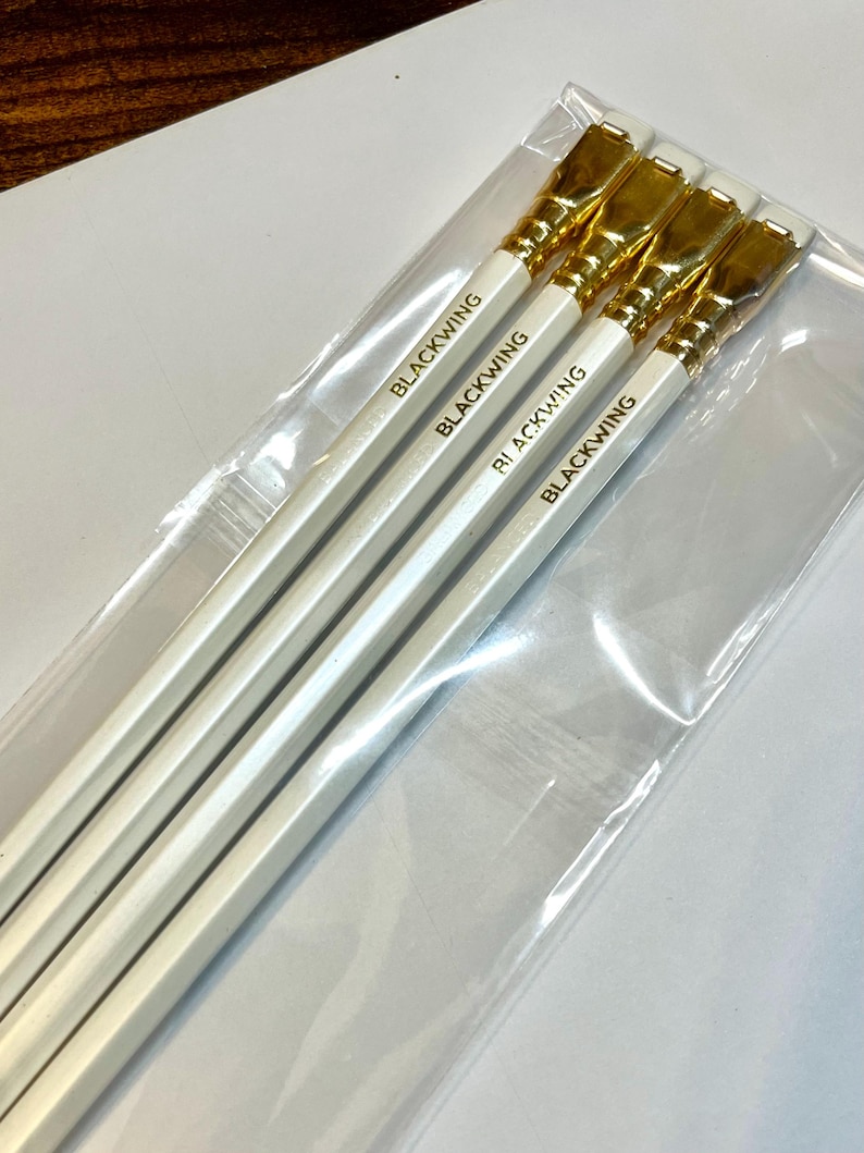 May include: Four white pencils with gold-colored erasers and the word "BLACKWING" printed in gold on each pencil.