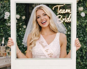 Op de afbeelding: Een vrouw lacht, omlijst door een groot bord met de tekst "Sophie's Hen Party". Ze draagt een sjerp met de tekst "Bride to Be" en een sluier. Het bord bevat ook de tekst "26 dagen tot ze ja zegt" en "28 juni 2026".