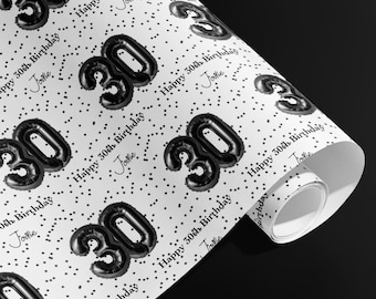 May include: White wrapping paper with a repeating pattern of black "30" balloon-style numbers and small black dots. The words "Happy 30th Birthday" and the name "Jodie" are also printed on the paper. The paper is rolled up.