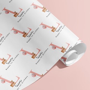 May include: White wrapping paper with a repeating pattern featuring the name "Laura", the text "Happy 1st birthday", and a pink letter "L" with a giraffe illustration. The paper is rolled up on a pink surface.