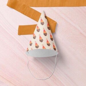 May include: A party hat with a white interior and a cone shape. The hat is decorated with a repeating pattern of faces wearing party hats. The background is a light pink surface with a mustard-colored ribbon.