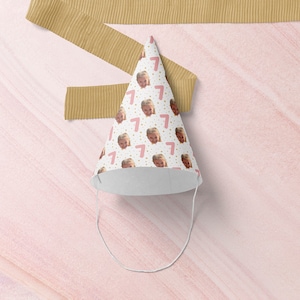 May include: A white party hat with a pink number seven and a child's face repeated on the surface. The hat has a white string attached to the top.