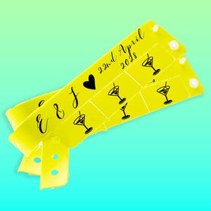 Pode incluir: Duas pulseiras amarelas com texto preto e gráficos de copos de cocktail. O texto inclui a data "22 de abril de 2028" e as iniciais "E & J" com um símbolo de coração. Cada pulseira tem fechos de botão brancos.