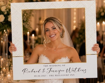 Op de afbeelding: Een witte fotobooth-frame met gouden geometrische accenten. Het frame heeft de tekst "THE WEDDING OF Richard & Jennifer Watkins 08.08.2027 | THE HILLCOURT INN". Een bruid houdt het frame vast en glimlacht.