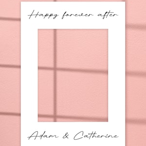 May include: White rectangular frame with the text "Happy forever after" at the top and "Adam & Catherine" at the bottom, set against a pink background. The frame casts a shadow.