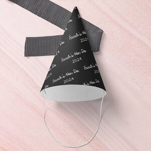 May include: Black party hat with white text that says "Sarah's Hen Do 2024" repeated on the hat. The hat is on a pink background.