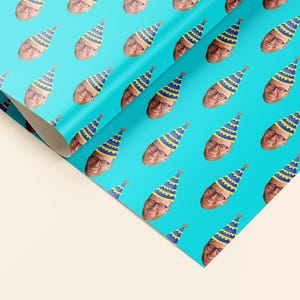 Könnte beinhalten: Eine Rolle türkisfarbenes Geschenkpapier mit einem sich wiederholenden Muster eines Gesichts mit Partyhut. Die Partyhüte sind blau und gelb. Das Papier ist teilweise abgerollt und zeigt die Rückseite.