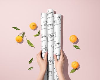 May include: Three rolls of white wrapping paper with a black and white pattern of the text "Simply Stationery" and the website address "simplystationery.etsy.com". The rolls are held in the hands of a person. The background is a light pink color with scattered orange mandarins and green leaves.