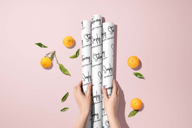 May include: Three rolls of white wrapping paper with a black and white pattern of the text "Simply Stationery" and the website address "simplystationery.etsy.com". The rolls are held in the hands of a person. The background is a light pink color with scattered orange mandarins and green leaves.