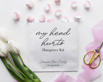 May include: White paper bag with the text "my head hurts Hangover Kit" and the text "Jessica's Hen Party 22/09/2022". The bag is surrounded by pink and white marshmallows, white tulips, and pink ribbon.