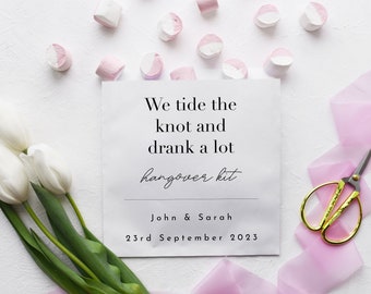 May include: White card with the text "We tide the knot and drank a lot hangover kit" with the names John & Sarah and the date 23rd September 2023. The card is surrounded by white tulips, pink ribbon, and pink and white marshmallows.