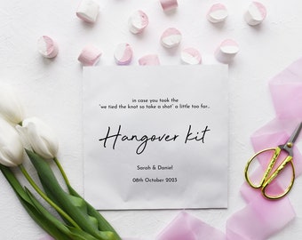 May include: White paper bag with the text "in case you took the 'we tied the knot so take a shot' a little too far... Hangover kit Sarah & Daniel 08th October 2023". The bag is surrounded by white tulips, pink marshmallows, and pink ribbon.