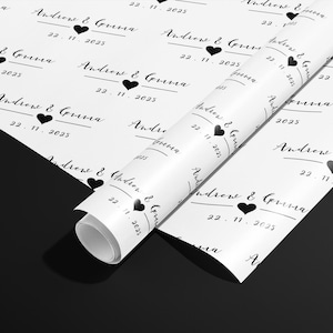 May include: White wrapping paper with a repeating pattern of the names "Andrew & Gemma", a black heart, and the date "22.11.2025". The paper is partially rolled, revealing the pattern.