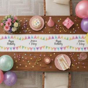May include: A birthday party table setting viewed from above. A white table runner with colourful pennants and the text "Happy Birthday" and "Jessica 7 Today!" is the focal point. Pink, purple, and green balloons, a cake, and gifts are also present.