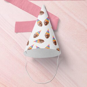 May include: A white party hat with a rainbow pattern and a repeating image of a child's face. The hat has an elastic band.
