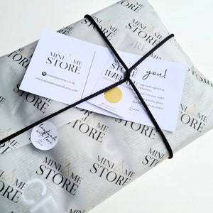 May include: A white gift bag with a black string tie. The bag is decorated with a repeating pattern of a tent with the words "Mini Me Store" printed on it. The bag is tied with a black string and has a small white sticker with the words "handmade with love" printed on it. There are two cards on top of the bag. One card says "Mini Me Store" and the other card says "Thank you!" and has a message thanking the customer for their order and supporting the small business.