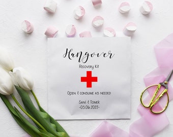 May include: White paper bag with the text "Hangover Recovery Kit" printed on it. A red cross symbol is below the text. The bag also includes the text "Open & consume as needed" and "Sani & Tomek -03.06.2022-."