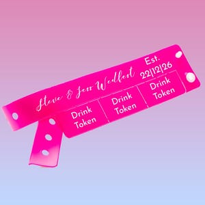 Pode incluir: Uma pulseira rosa brilhante com o texto "Steve & Jess Wedfest" em itálico. A pulseira tem três seções rotuladas como "Drink Token" e a data "22|12|26". Possui um fecho de pressão.