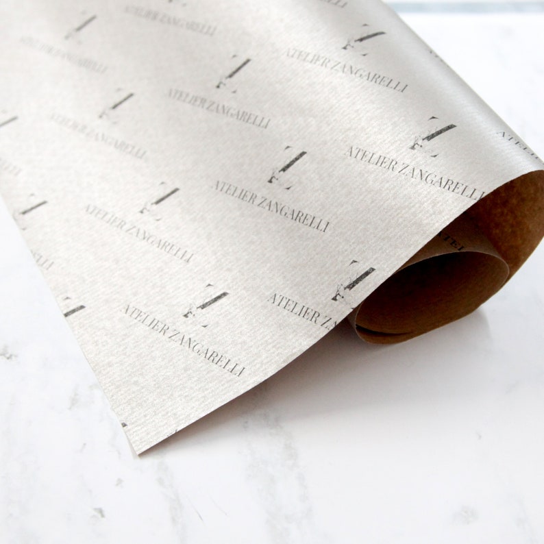 K&ouml;nnte beinhalten: Eine Rolle silbernes Geschenkpapier mit einem sich wiederholenden Muster des Textes "Atelier Zangarelli" in Schwarz.